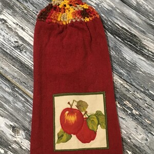 May include: A red kitchen towel with a crocheted top in shades of brown, yellow, and orange. The towel features an embroidered square with two red apples and green leaves. The towel is designed for the kitchen.
