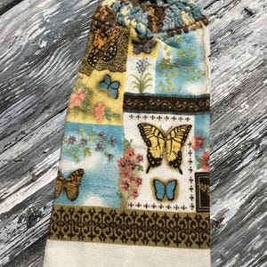 May include: A decorative kitchen towel with a crocheted top. The towel features a patchwork design with butterflies, flowers, and a brown decorative border. The color palette includes blue, yellow, and brown.