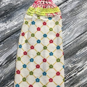 May include: A white kitchen towel with a floral pattern in red, blue, and green, connected by yellow lines. The top features a crocheted loop in pink, yellow, and blue, with a pink button closure. The towel is designed for hanging.