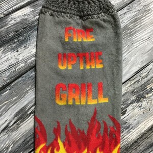 May include: Gray kitchen towel with a crocheted top and button closure. The towel features the text "FIRE UP THE GRILL" in orange and yellow, with a flame design at the bottom. The top is gray with yellow and orange accents.