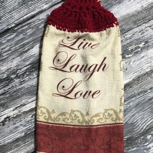 Pode incluir: Uma toalha de cozinha creme com as palavras "Live Laugh Love" em escrita bordô. A parte superior é um design de crochê bordô com uma alça creme para pendurar. Uma borda estampada bordô e dourada acentua a parte inferior.