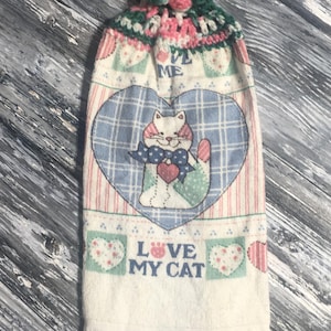 May include: A decorative kitchen towel with a crocheted top in green, pink, and white. The towel features a cat design inside a blue heart, with the text "LOVE MY CAT".