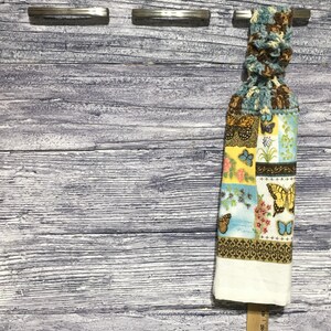 Peut inclure: Un torchon de cuisine d&eacute;coratif avec un dessus en crochet et un bas &agrave; motifs avec des papillons et des motifs floraux. Le torchon est blanc en bas et est suspendu &agrave; une barre m&eacute;tallique. L'arri&egrave;re-plan est une surface en bois vieilli.