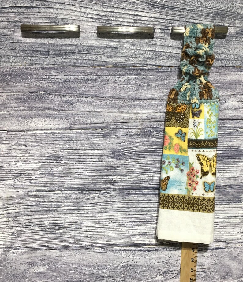 Peut inclure: Un torchon de cuisine d&eacute;coratif avec un dessus en crochet et un bas &agrave; motifs avec des papillons et des motifs floraux. Le torchon est blanc en bas et est suspendu &agrave; une barre m&eacute;tallique. L'arri&egrave;re-plan est une surface en bois vieilli.