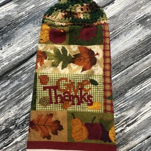 May include: A fall-themed hanging kitchen towel with a crocheted top and button closure. The towel features a patchwork design with autumn leaves, apples, pears, and the words "Give Thanks". The bottom is a cream-coloured towel.