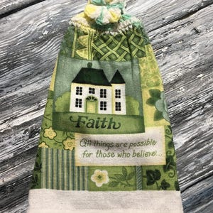 May include: A green and white hanging kitchen towel with a crocheted top. The towel features a house graphic with the word "Faith" and the text "All things are possible for those who believe..."