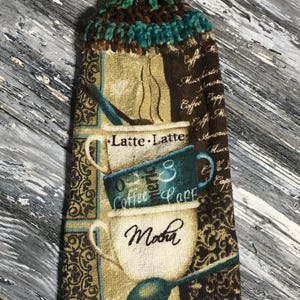 May include: A decorative kitchen towel with a crocheted top and a button closure. The towel features coffee-themed graphics, including coffee cups and the words "Latte Latte" and "Mocha". The color scheme includes brown, teal, and beige.