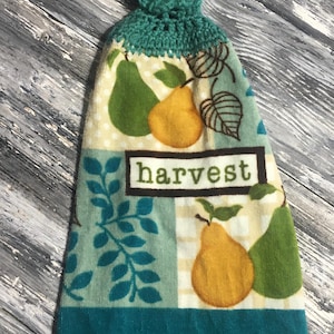 May include: A teal and cream-colored kitchen towel with a crocheted top. The towel features a "Harvest" text design and a pattern of green and yellow pears. The bottom edge is a solid teal color.