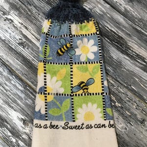 May include: A hanging kitchen towel with a blue crocheted top and a patterned bottom. The bottom features a grid design with bees, daisies, and the text "as a bee - Sweet as can be". The towel has a white, fringed edge.