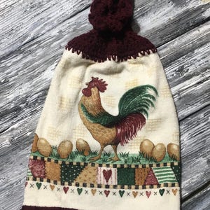 Könnte beinhalten: Ein dekoratives Küchentuch mit einem Hahn-Design. Das Tuch hat einen cremefarbenen Hintergrund mit einer farbenfrohen Hahn-Illustration, Eiern und einem Patchwork-Rand. Die Oberseite ist eine bordeauxfarbene Häkelschlaufe.