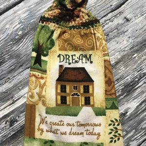 May include: A decorative kitchen towel with a crocheted top. The towel features a house illustration with the word "DREAM" above it and the quote "We create our tomorrows by what we dream today."