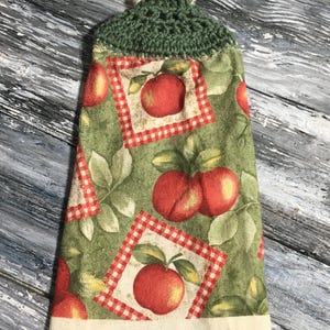 May include: A kitchen towel with a green crocheted top and a button closure. The towel features a pattern of red apples, green leaves, and red and white checkered squares on a green background. The bottom edge is a cream color.