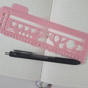 May include: A pink plastic stencil ruler with various shapes and a ruler edge, alongside a black pen, rests on an open notebook with dotted pages. The stencil features stars, circles, and other geometric designs, ideal for journaling and crafting.