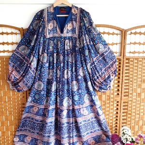 May include: A blue dress with a V-neck and a floral pattern in shades of blue, pink, and white. The dress has long sleeves and a loose fit. The dress is hanging on a wooden hanger.