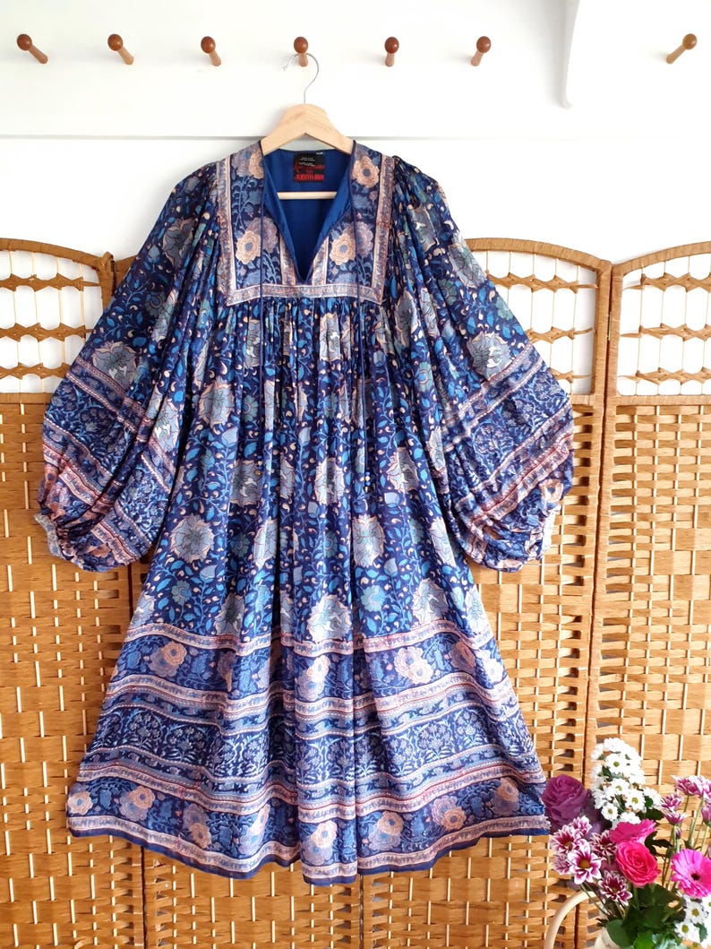 May include: A blue dress with a V-neck and a floral pattern in shades of blue, pink, and white. The dress has long sleeves and a loose fit. The dress is hanging on a wooden hanger.
