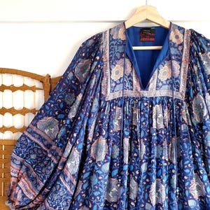 May include: A blue floral dress with long sleeves, hanging on a wooden hanger. The dress has a V-neck and is patterned with blue, light blue, and beige floral motifs. The dress's fabric is soft and flowing.
