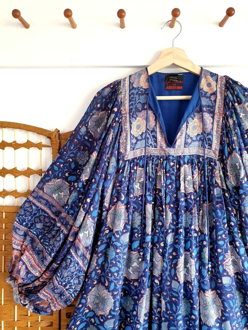 May include: A blue floral dress with long sleeves, hanging on a wooden hanger. The dress has a V-neck and is patterned with blue, light blue, and beige floral motifs. The dress's fabric is soft and flowing.