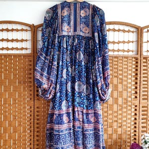 May include: A long, blue dress with a floral design, hanging on a wooden hanger. The dress features long sleeves and a detailed pattern. The background includes a wooden folding screen and a white wall with wooden pegs.