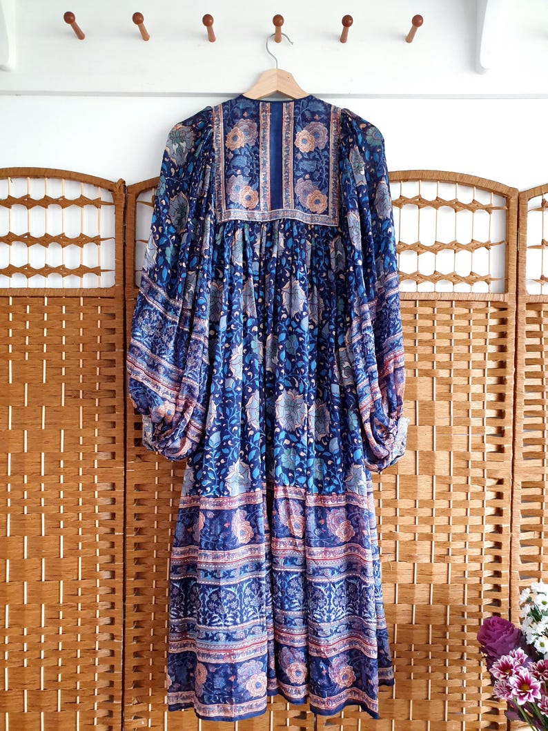 May include: A long, blue dress with a floral design, hanging on a wooden hanger. The dress features long sleeves and a detailed pattern. The background includes a wooden folding screen and a white wall with wooden pegs.