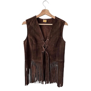 Puede incluir: Un chaleco de ante marrón con cuello en V y flecos. El chaleco tiene un cierre de cordones y está colgado de una percha de madera. El chaleco es de color marrón medio.