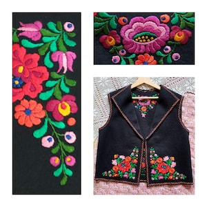 May include: A black vest with colorful floral embroidery. The embroidery features red, pink, yellow, and blue flowers with green leaves. The vest has a collar and is displayed on a wooden hanger.