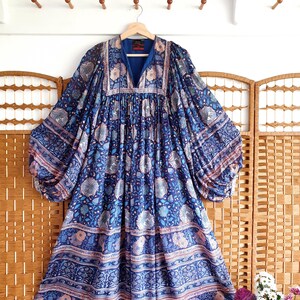 May include: A blue patterned dress with long, voluminous sleeves and a V-neck. The dress features a floral design in shades of blue, pink, and white. The dress is hanging on a wooden hanger.