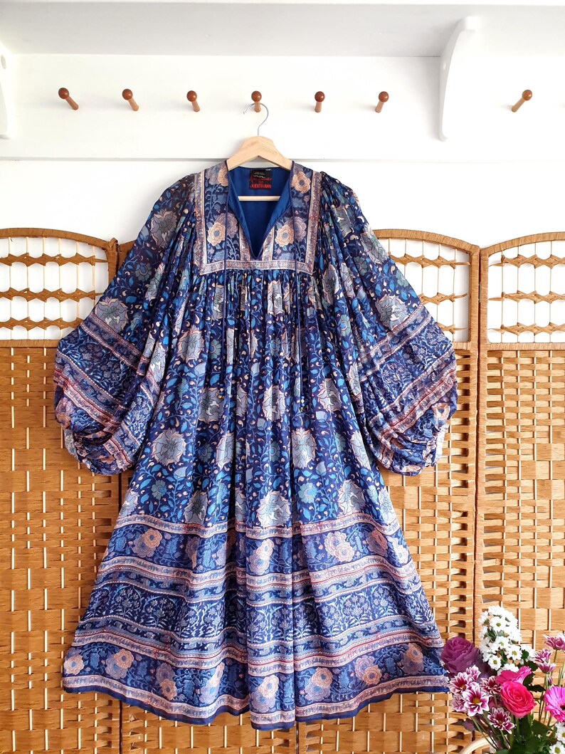 May include: A blue patterned dress with long, voluminous sleeves and a V-neck. The dress features a floral design in shades of blue, pink, and white. The dress is hanging on a wooden hanger.