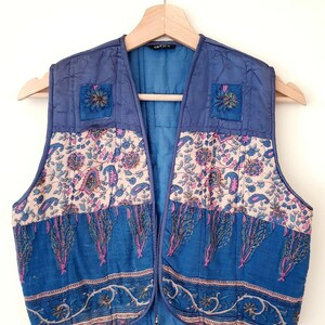 May include: A blue quilted vest with floral and paisley patterns. The vest has a zip closure and is hanging on a wooden hanger. The vest features a patchwork design with blue, cream, and pink colors. The vest has two small floral patches on the upper chest area.