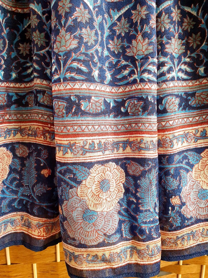 May include: Close-up of a fabric with a floral pattern in navy blue, teal, and cream. The design features large flowers and intricate leaf patterns, with borders of geometric and floral motifs.
