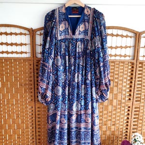 May include: A long, blue dress with a floral pattern and long sleeves hangs on a wooden hanger. The dress features a V-neck and a mix of blue, pink, and white floral designs. The dress is made of a lightweight fabric.