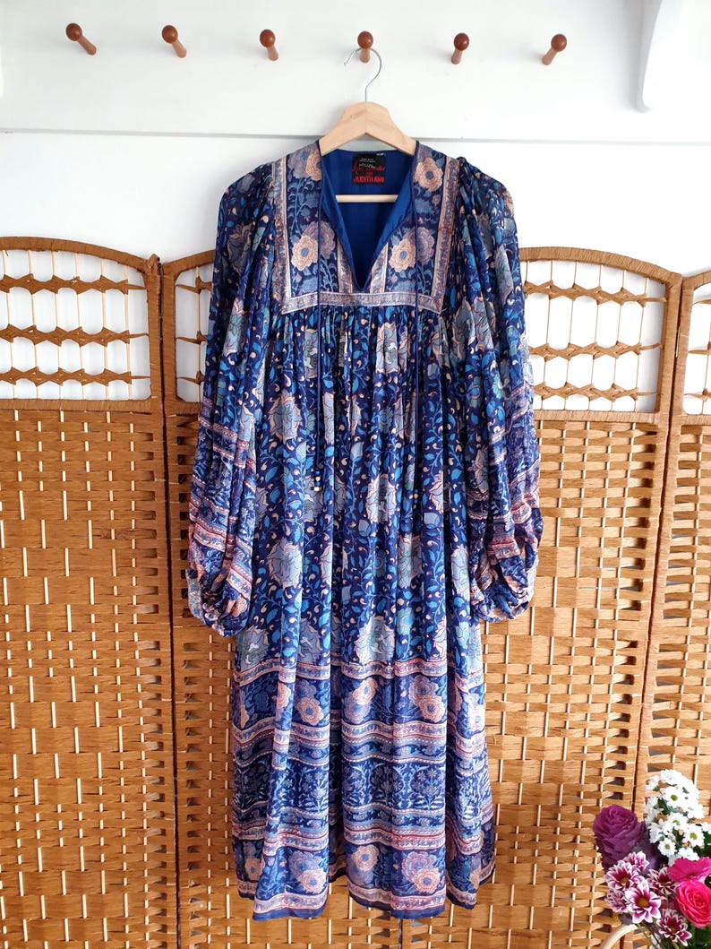 May include: A long, blue dress with a floral pattern and long sleeves hangs on a wooden hanger. The dress features a V-neck and a mix of blue, pink, and white floral designs. The dress is made of a lightweight fabric.