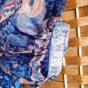 May include: Close-up of a blue fabric with a floral pattern in shades of blue, red, and beige. The fabric has a detailed design with a border featuring a light blue floral motif. The fabric is draped over a wooden surface.
