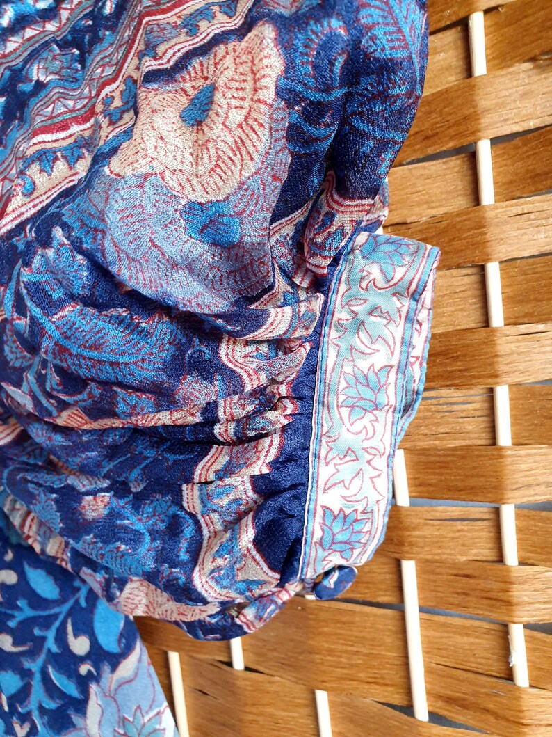 May include: Close-up of a blue fabric with a floral pattern in shades of blue, red, and beige. The fabric has a detailed design with a border featuring a light blue floral motif. The fabric is draped over a wooden surface.