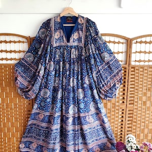 May include: A blue patterned dress with long sleeves, hanging on a wooden hanger. The dress features a V-neck and a floral design. The background includes a wooden screen and a white wall with wooden pegs.