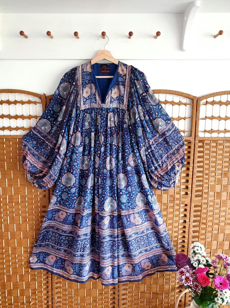 May include: A blue patterned dress with long sleeves, hanging on a wooden hanger. The dress features a V-neck and a floral design. The background includes a wooden screen and a white wall with wooden pegs.