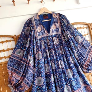May include: A long blue dress with a V-neck and floral print. The dress has long, wide sleeves and hangs on a wooden hanger. The floral pattern incorporates shades of blue, peach, and white, creating a bohemian style.