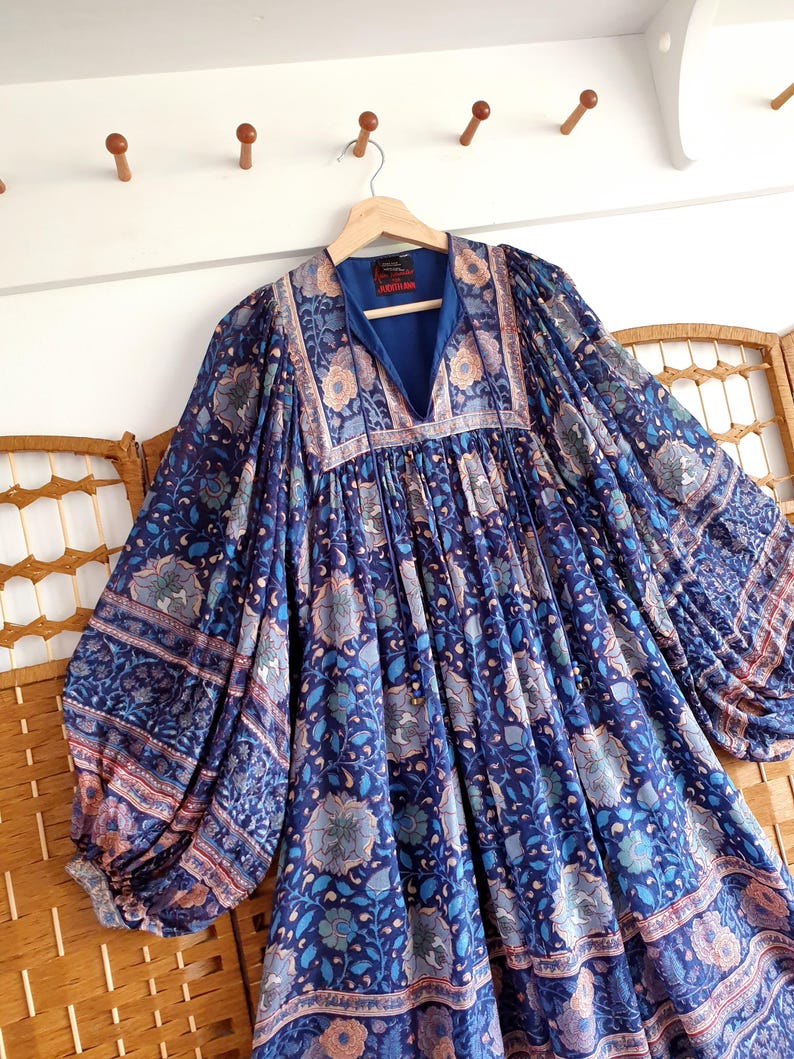 May include: A long blue dress with a V-neck and floral print. The dress has long, wide sleeves and hangs on a wooden hanger. The floral pattern incorporates shades of blue, peach, and white, creating a bohemian style.