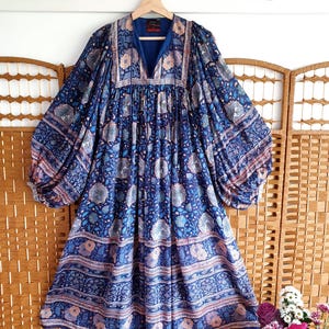 May include: A long blue dress with a floral pattern, hanging on a wooden hanger. The dress has a V-neck and a flowing, bohemian style. The fabric is decorated with floral and geometric designs in shades of blue, pink, and white. The dress has long sleeves.