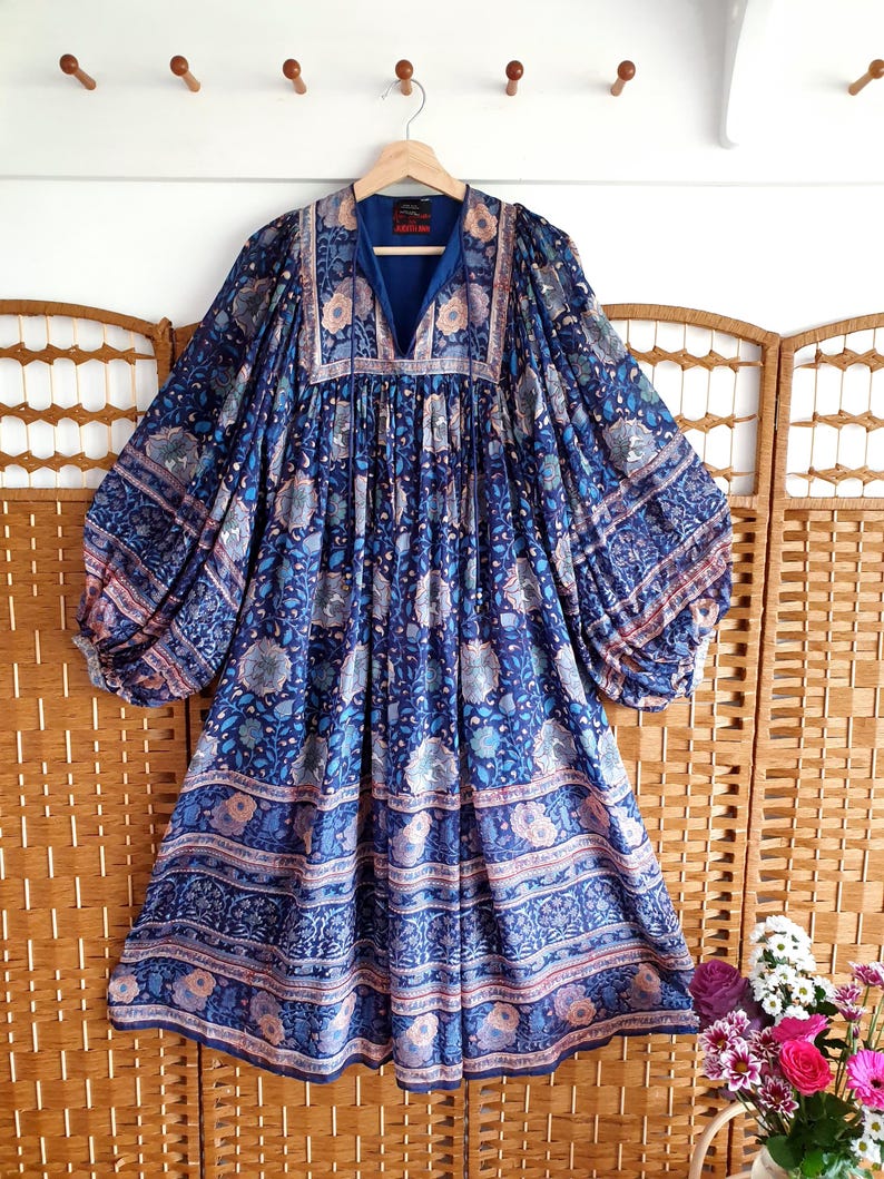 May include: A long blue dress with a floral pattern, hanging on a wooden hanger. The dress has a V-neck and a flowing, bohemian style. The fabric is decorated with floral and geometric designs in shades of blue, pink, and white. The dress has long sleeves.