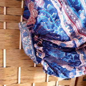 May include: Close-up of patterned fabric in shades of blue, red, and white. The fabric displays a floral and geometric design. The fabric is draped over a woven wooden structure. The pattern is intricate and detailed.