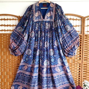 May include: A long, blue floral dress with a V-neck and voluminous sleeves. The dress has a tiered skirt and is patterned with flowers in blue, pink, and white. The dress is hanging on a wooden hanger, showcasing its flowing design.