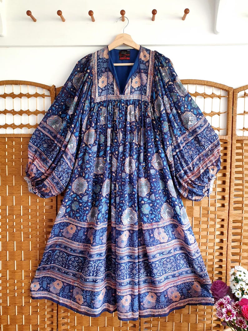 May include: A long, blue floral dress with a V-neck and voluminous sleeves. The dress has a tiered skirt and is patterned with flowers in blue, pink, and white. The dress is hanging on a wooden hanger, showcasing its flowing design.