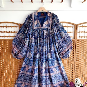 May include: A blue patterned dress with long sleeves and a V-neck. The dress has a floral design in shades of blue, pink, and white. It is hanging on a wooden hanger. The dress has a flowing, loose fit.