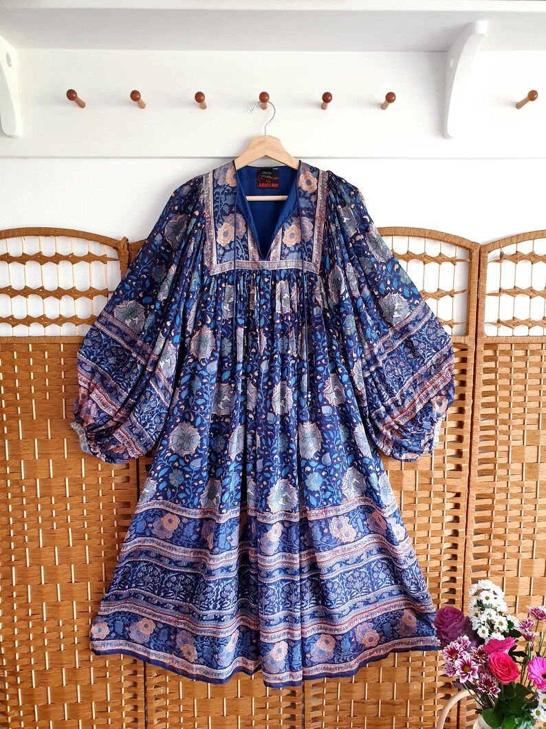 May include: A blue patterned dress with long sleeves and a V-neck. The dress has a floral design in shades of blue, pink, and white. It is hanging on a wooden hanger. The dress has a flowing, loose fit.