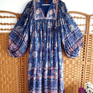 May include: A long, flowing dress in a deep blue hue, adorned with a floral pattern in pink, white, and blue. It has a V-neck and long, balloon sleeves. The dress is suspended from a wooden hanger, showcasing its intricate design.