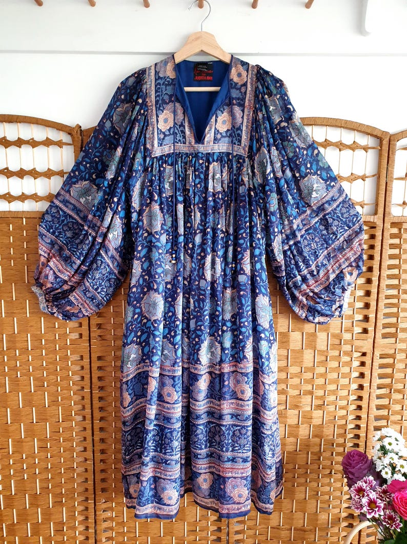 May include: A long, flowing dress in a deep blue hue, adorned with a floral pattern in pink, white, and blue. It has a V-neck and long, balloon sleeves. The dress is suspended from a wooden hanger, showcasing its intricate design.