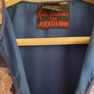 May include: Close-up of a blue garment with a floral pattern in red and cream. The garment has a dark blue lining and a label that reads "Ritu Kumar for JUDITH ANN" in red text.