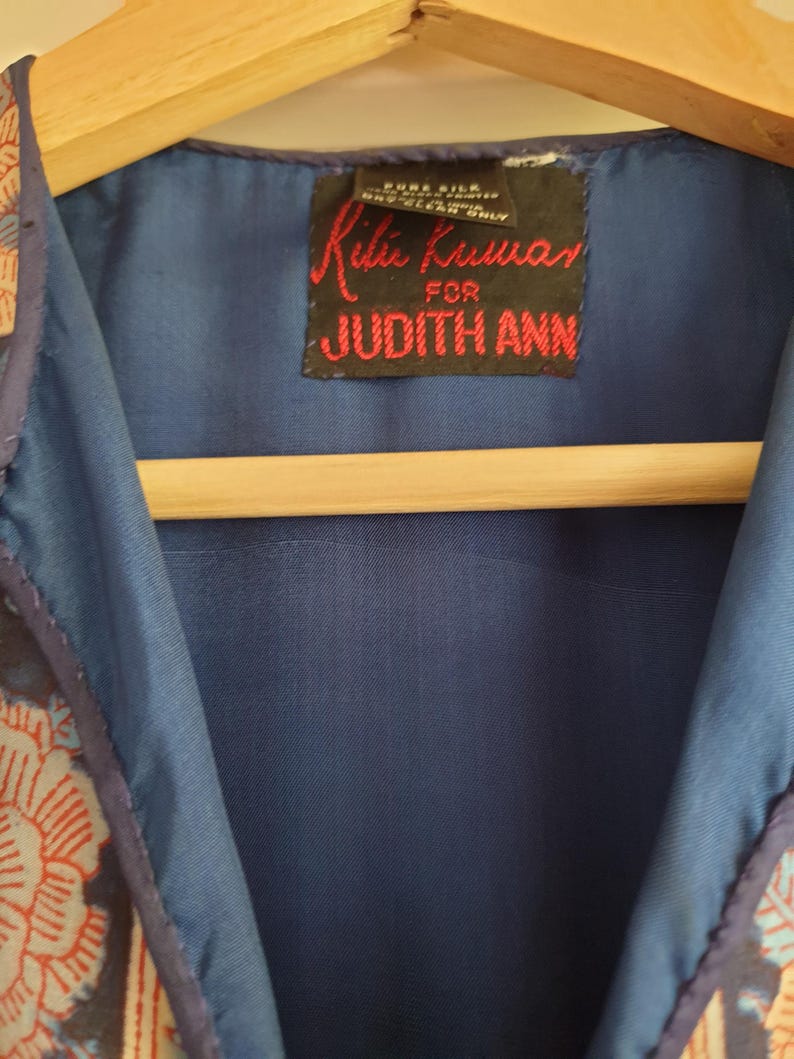 May include: Close-up of a blue garment with a floral pattern in red and cream. The garment has a dark blue lining and a label that reads "Ritu Kumar for JUDITH ANN" in red text.