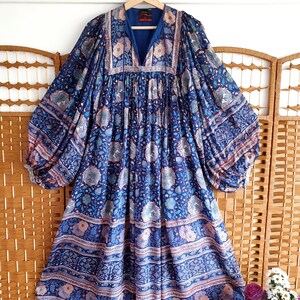 May include: A long-sleeved blue dress with a V-neck and floral design. The dress has a flowing shape with wide sleeves and a patterned border. The fabric features blue, light blue, and peach tones.