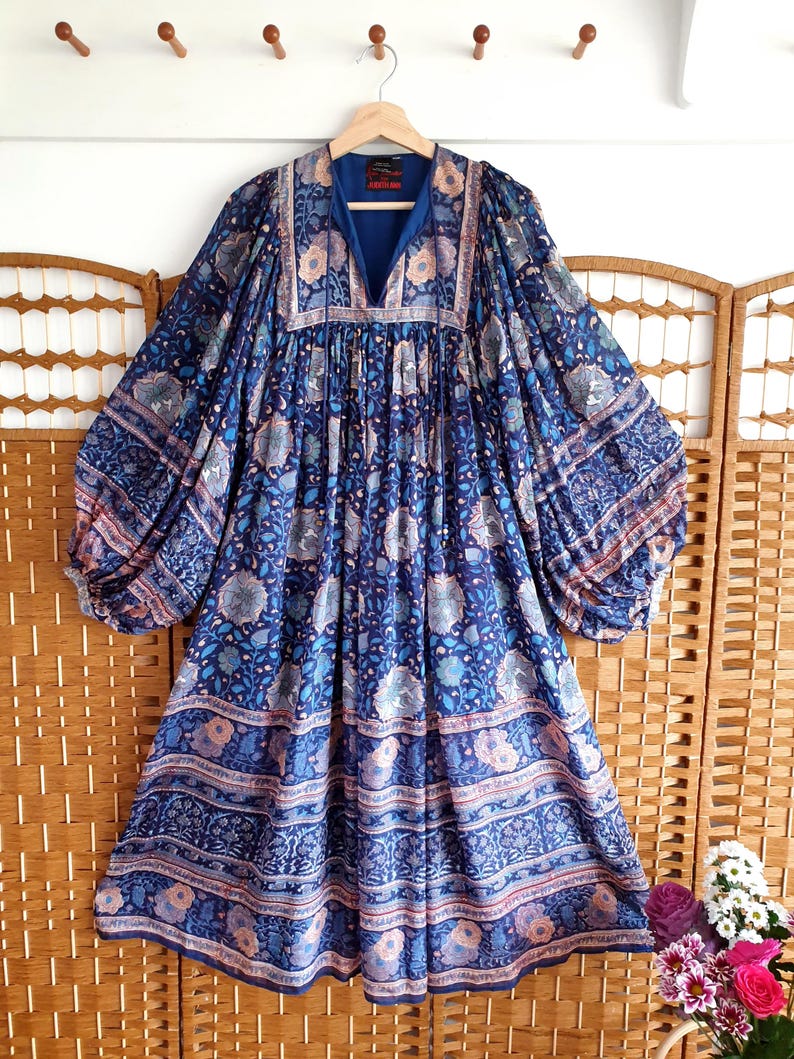 May include: A long-sleeved blue dress with a V-neck and floral design. The dress has a flowing shape with wide sleeves and a patterned border. The fabric features blue, light blue, and peach tones.